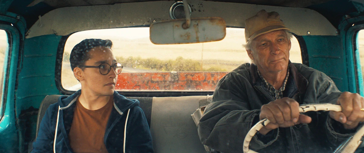 A young man on the left and older man on the right are seated in the passenger and driver seats of an old truck, looking at each other.